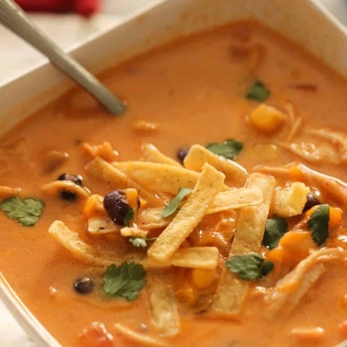 A bowl of creamy tortilla soup with tortilla strips, black beans, corn, cilantro, and a spoon.