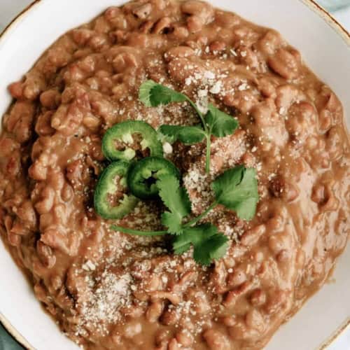 A bowl of refried beans topped with sliced jalapeños, cilantro, and grated cheese, placed on a blue cloth.